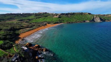 Gozo 'daki en güzel Malta sahilinin hava aracı görüntüsü, berrak mavi suyu ve yemyeşil bitki örtüsüyle büyüleyici dalgaları olan canlı kırmızı Ramla plajı ve güneşli bir günde kont sabahında ünlü Mixta Mağarası arka planı.