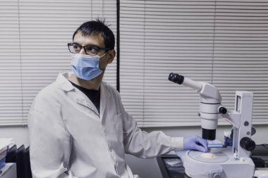 Laboratuvarında mikroskop kullanarak yüz maskesi takan erkek bilim adamının stok fotoğrafı..