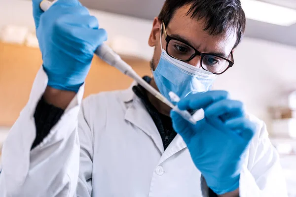 Laboratuvarında yüz maskesi takan erkek bilim adamının test tüplerini doldururken çekilmiş fotoğrafı..