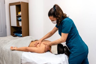 Yüz maskesi takan fizyoterapistin hastaya sedyede yatarken masaj yaptığı fotoğraf..