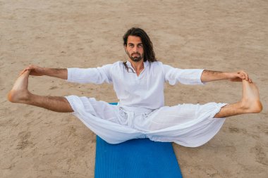 Uzun saçlı yetişkin bir adamın plajda yoga pozu verirken çekilmiş resmi..