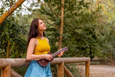 Genç beyaz kadın parkta ukelele oynuyor.