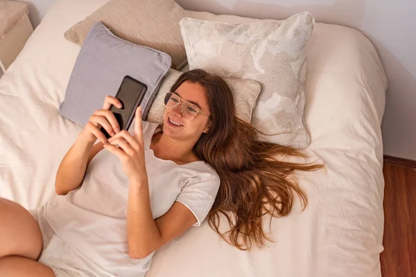 Młoda Latynoska z telefonem komórkowym leżąca w łóżku — Zdjęcie stockowe