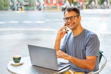 Genç Latin adam dışarıda cep telefonu ve bilgisayarla mutlu bir şekilde çalışıyor..