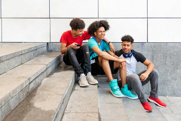 Üç afro Latin erkek arkadaş grubu merdivenlerde oturup cep telefonu kullanarak eğleniyor. Boşluğu kopyala.