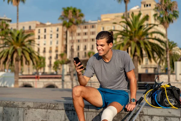 Young latin man with crutches using mobile phone after doing exercise