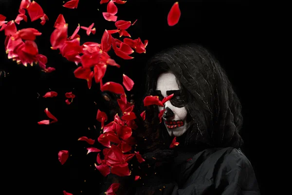 woman with a make-up skeleton stands in black clothes and a transparent hood and throws up red roses on a black background