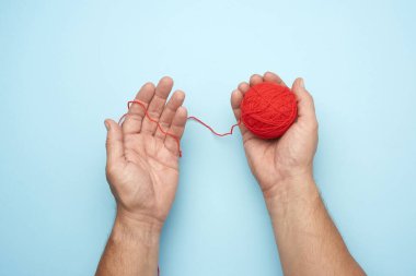 İki erkek el, kırmızı yün bir iplik topunu tutuyor, mavi arka plan, üst manzara.