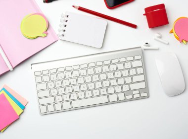 Beyaz masaüstünde beyaz kablosuz klavye, bilgisayar faresi ve kağıt defter, üst görünüm. İşyeri