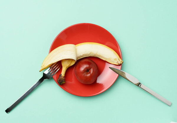 red ripe apple and banana lie in a red round ceramic plate, near a metal knife and fork, top view