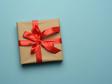 Kahverengi kağıda sarılı bir kutu ve kırmızı ipek kurdeleyle bağlanmış bir fiyonk, mavi arka planda hediye, üst manzara, kopyalama alanı