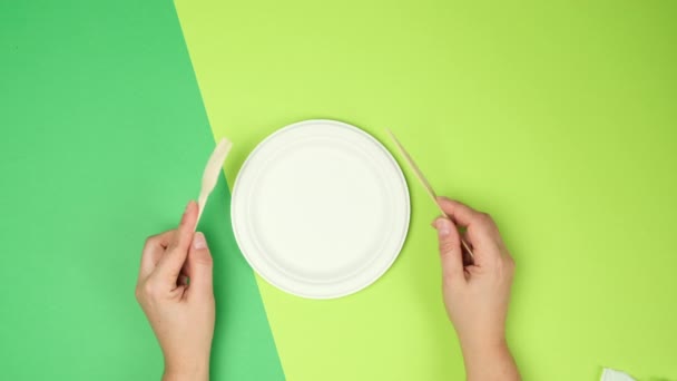空の白い紙皿と女性の手は緑の背景に使い捨てのフォークとナイフを持っていますトップビュー — ストック動画