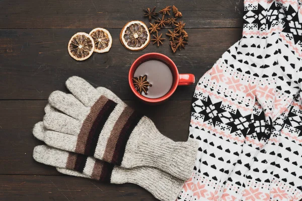 gray knitted mittens and red cup with a drink on a brown wooden table, top view