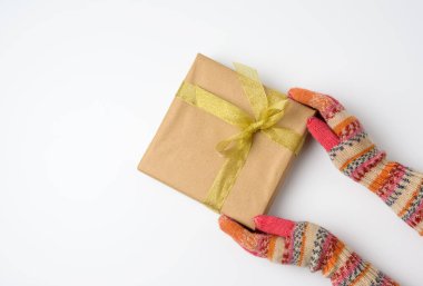 Örgü eldivenli kadın elleri kahverengi kağıda sarılı kare bir kutu tutuyor ve beyaz arka planda altın bir kurdele ile bağlanıyor.