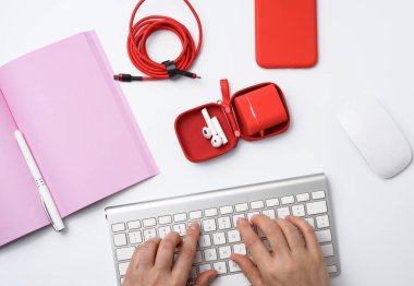 İki kadın eli ve bir beyaz klavye, açık bir defter, kulaklık, kırmızı akıllı telefon, beyaz masa, üst görünümü olan serbest çalışan işyeri.