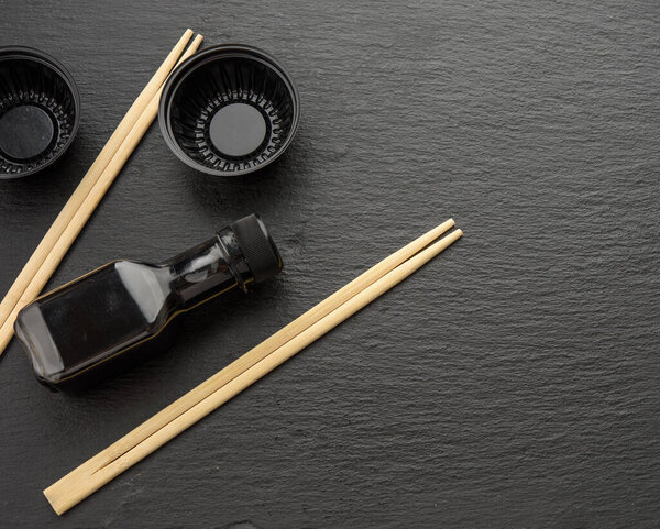 wooden sticks for sushi, bottle of soy sauce and disposable plastic plates on black background, utensils for delivery