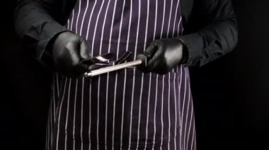 Çizgili önlüklü ve siyah üniformalı bir adam metal bileyicili bir mutfak bıçağını keskinleştiriyor. 