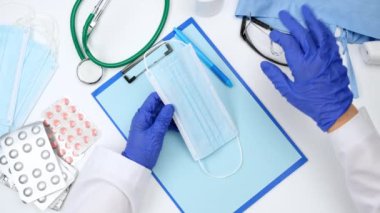 Mavi letax eldivenli iki kadın eli, doktorun iş yerini gösteren tek kullanımlık bir tıbbi yüz maskesi taşıyor.