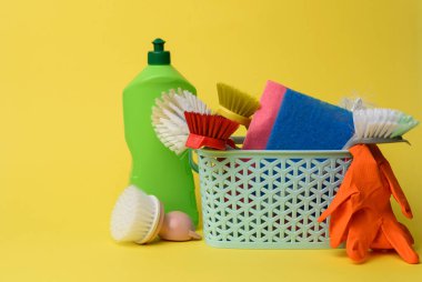 Temizlik için mavi plastik sepet, fırçalar, süngerler ve lastik eldivenler, sarı arka plan.