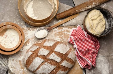 Masanın üzerinde bir somun ekmek ve malzemeler, mutfak gereçleri yakınlarda, üst tarafta.