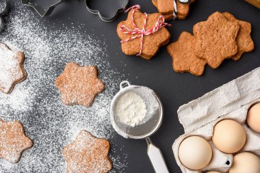 Yıldız şeklinde fırınlanmış zencefilli kurabiyeler üzerine pudra şekeri serpiştirilmiş siyah bir masa, üst manzara
