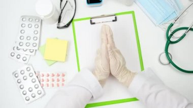 Doktorun masasının üst görüntüsü, dua pozisyonunda eğilmiş, beyaz lateks eldivenler giyiyor. 