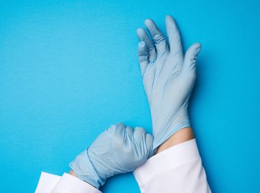 Beyaz önlüklü kadın doktor ellerine mavi lateks steril eldiven takıyor, mavi arka plan, yakın plan.