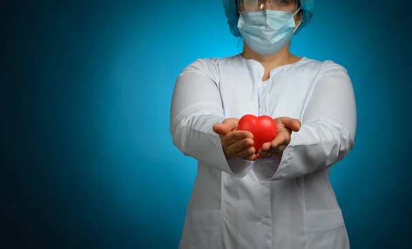 female medic in a white coat, a mask stands and holds a red heart on a blue background, the concept of donation and kindness