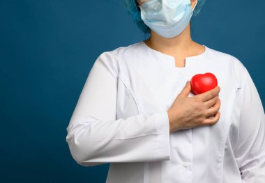 Beyaz önlüklü kadın doktor, maske duruyor ve mavi arka planda kırmızı bir kalp taşıyor, bağışta bulunma ve şefkat kavramı.