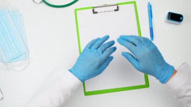 Doktorun masasının üst görüntüsü, dua pozisyonu almış, mavi lateks eldivenler giymiş ve başında koruyucu bir başlık var.