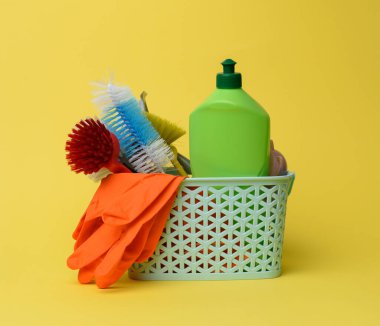Temizlik için mavi plastik sepet, fırçalar, süngerler ve lastik eldivenler, sarı arka plan.