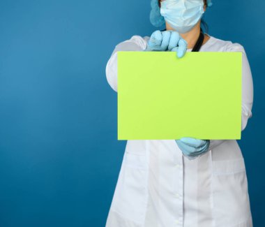 Beyaz bir doktor, tek kullanımlık bir maske, koruyucu plastik bardaklar ve mavi arka planda duran bir şapka ve boş yeşil bir sayfa tutuyor.
