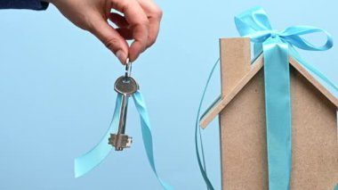 Kadın eli, model bir evin arka planında mavi kurdeleli metal bir anahtar tutuyor. Emlak satın alma konsepti, ipotek, mavi özgeçmiş