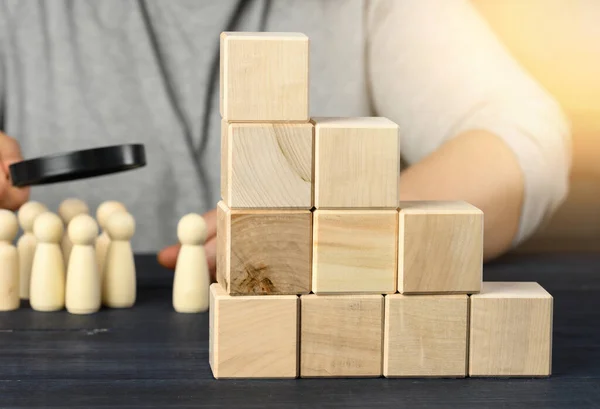 ladder of wooden cubes on a steel, behind a man under a magnifying glass examines wooden figures. Recruitment concept, search for talented employees, career advancement