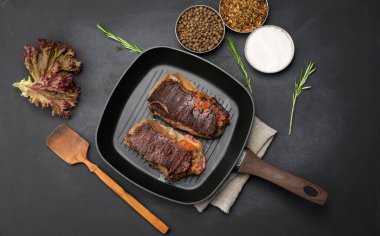 İki kızarmış et parçası siyah bir masada tavada, baharatların yanında, yemeğin hazır olma derecesi iyi.