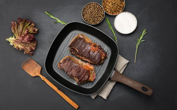 İki kızarmış et parçası siyah bir masada tavada, baharatların yanında, yemeğin hazır olma derecesi iyi.