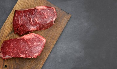 Kahverengi ahşap bir tahtada iki çiğ biftek, siyah masalar, üst manzara.