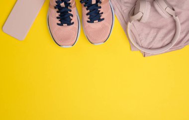 Bir çift pembe spor ayakkabı, kablosuz kulaklık, bir akıllı telefon ve sarı arka planda akıllı bir saat, yazıtlar için bir yer, düz uzanma