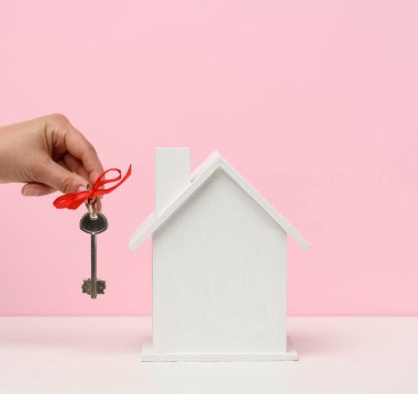 Anahtarları ve pembe arka planda ahşap bir evi tutan el. Emlak kiralama, alım ve satım konsepti. Emlakçılık, bina onarımı ve bakımı, kopyalama alanı