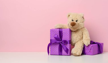 Kağıda sarılı bir kutu ve beyaz masada kırmızı ipek kurdele tutan sevimli kahverengi ayıcık. Ödül ve tebrikler, pembe arka plan, kopyalama alanı