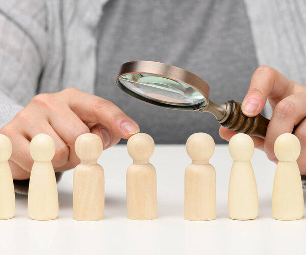 figurines of men on a white table, a female hand holds a magnifying glass over one. concept of searching for employees in the company, recruiting personnel, identifying talented and strong personalities