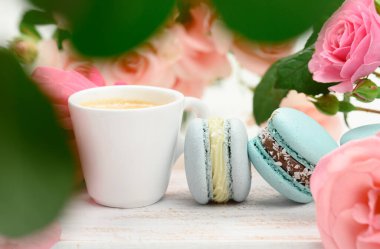 Beyaz bir masanın üzerinde kahve ve makarna ile beyaz seramik fincan, bir buket pembe gülün arkasında, yakın