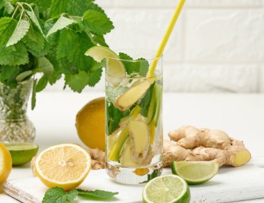 Limonlu şeffaf cam bardak, kireç parçaları ve nane yaprakları ve beyaz bir masada zencefil parçaları, detoks.