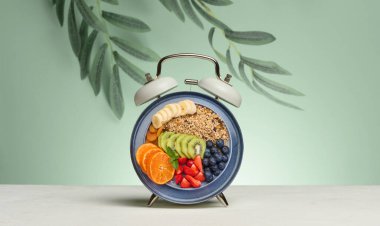 Alarm clock with a dial replaced by a plate full of a healthy breakfast. The plate contains granola, banana slices, kiwi, tangerine, strawberries, and blueberries. Green background 