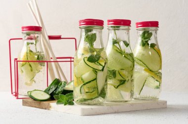 Kırmızı kapaklı cam şişeler, içinde salatalık, limon dilimleri ve nane yaprakları bulunan detoks suyla dolu beyaz masa.