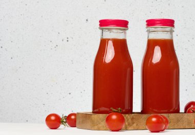 Kırmızı başlıklı domates suyu ile dolu cam şişeler. Taze kırmızı domatesler ahşap bir standın üzerinde ve önlerinde beyaz bir zemin var.