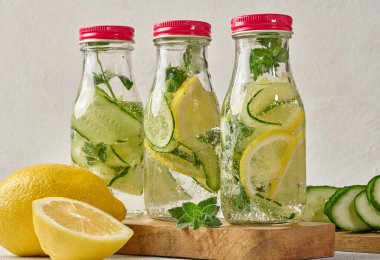 Kırmızı kapaklı cam şişeler, içinde salatalık, limon dilimleri ve nane yaprakları bulunan detoks suyla dolu beyaz masa.