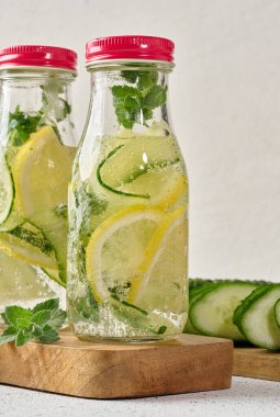 Kırmızı kapaklı cam şişeler, içinde salatalık, limon dilimleri ve nane yaprakları bulunan detoks suyla dolu beyaz masa.