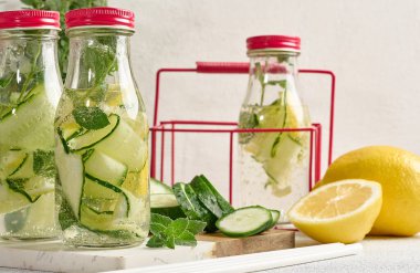 Kırmızı kapaklı cam şişeler, içinde salatalık, limon dilimleri ve nane yaprakları bulunan detoks suyla dolu beyaz masa.