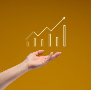 Bir kadının eli, yukarı doğru eğilimli, bar tablosu ve sarı arka planda büyüme çizgisi olan parıldayan dijital bir grafiği tutar. İş büyümesi ve başarı kavramı.